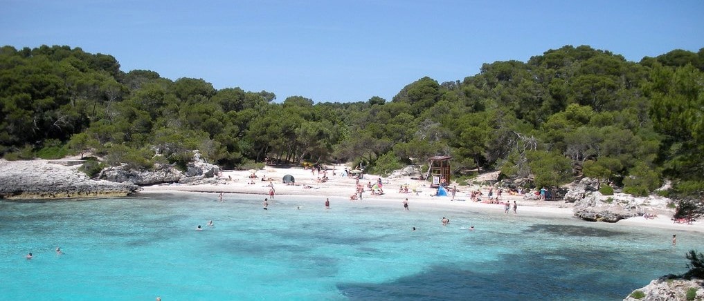 Plage de sable blanc à Minorque avec au premier plan la mer avec une eau turquoise et cristalline et à l'arrière plan la forêt.